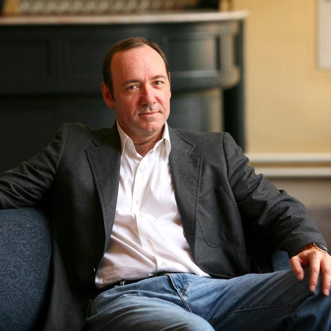A man in a gray blazer and white shirt sits casually on a blue sofa, resting one arm on the backrest. The background features a dark cabinet and a softly lit room.