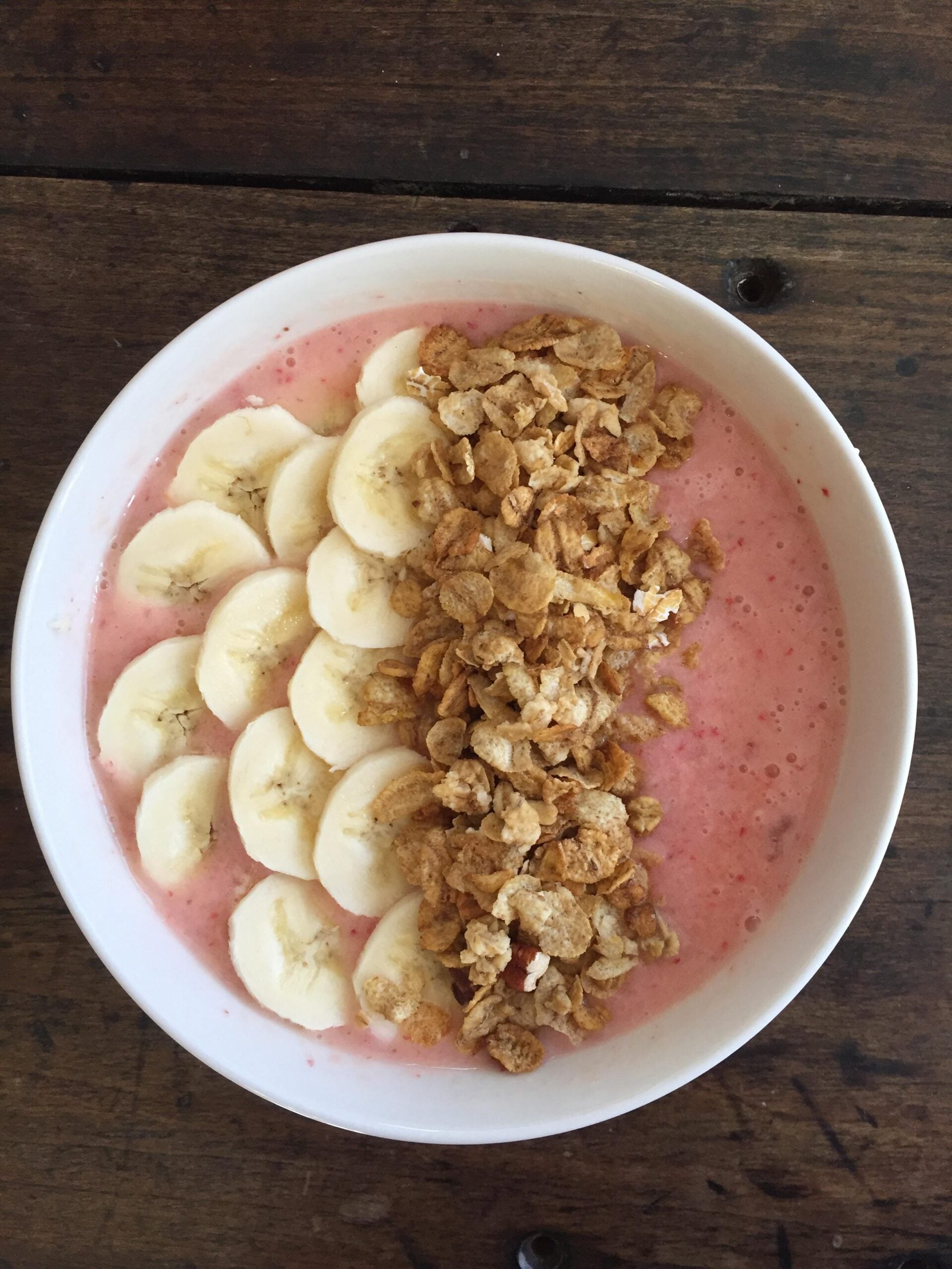 A white bowl filled with a pink smoothie, topped with sliced bananas on one side and granola on the other, placed on a rustic wooden table.