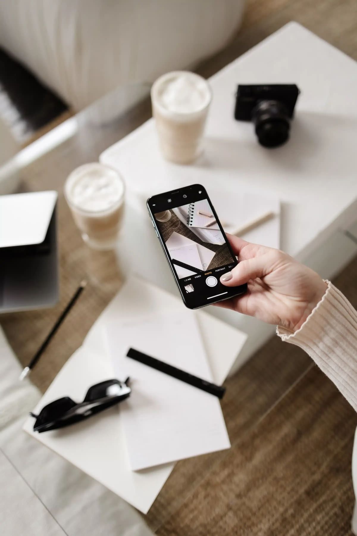 A person holding a smartphone takes a photo of two coffee cups on a table, surrounded by a camera, sunglasses, pen, notebook, laptop, and papers.