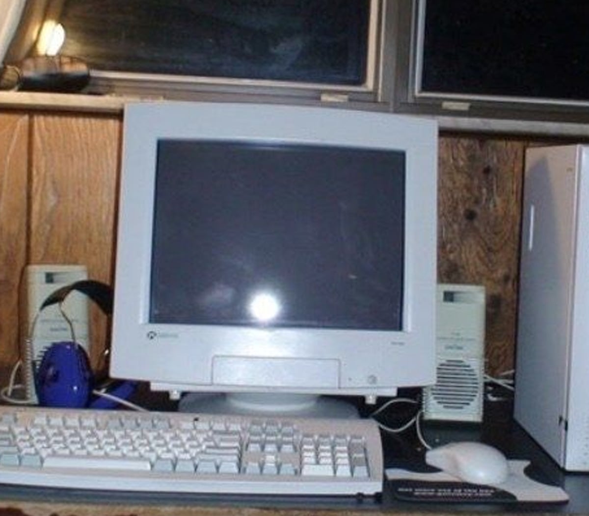 A vintage desktop computer setup with a CRT monitor, white keyboard, two speakers, a mouse on a mouse pad, and blue headphones, all placed on a wooden desk in front of a wooden wall.