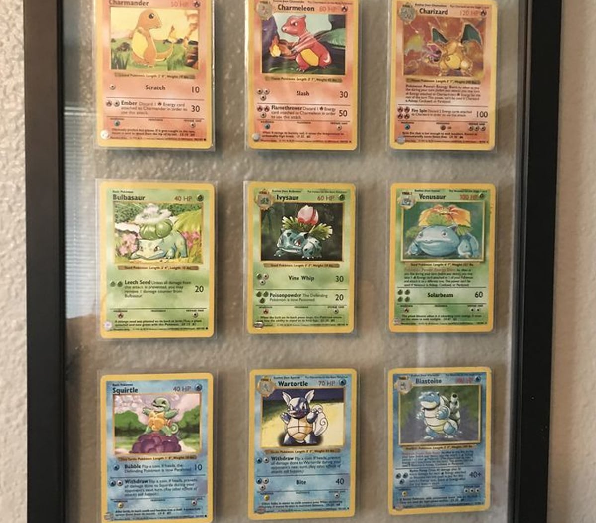 A framed display of nine Pokémon trading cards, featuring the evolutionary lines of Charmander, Bulbasaur, and Squirtle, arranged in three rows and three columns against a beige wall.