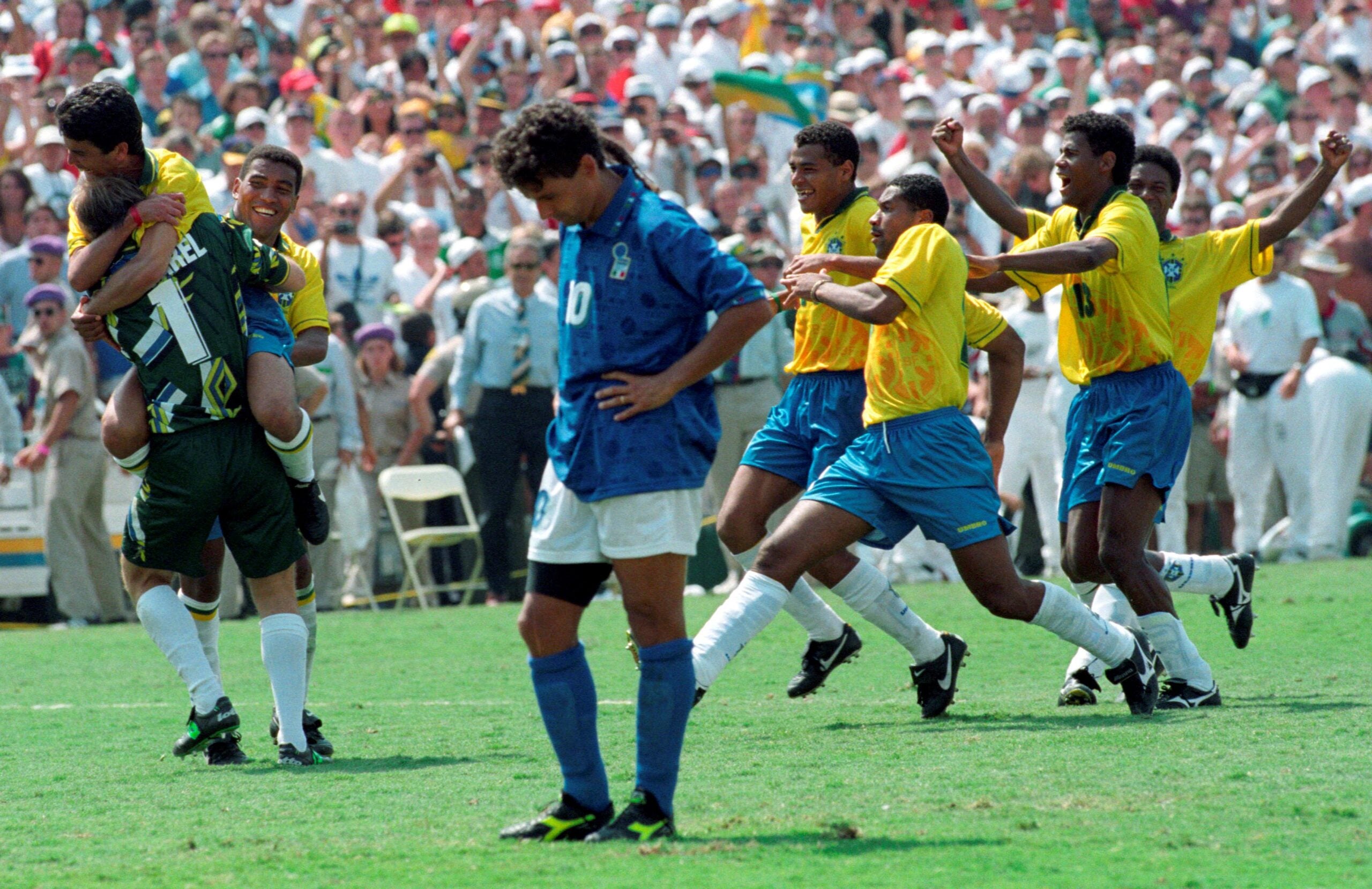 Brazilian soccer players in yellow jerseys celebrate on the field as an Italian player in blue stands with his head down, while a crowd watches in the background.