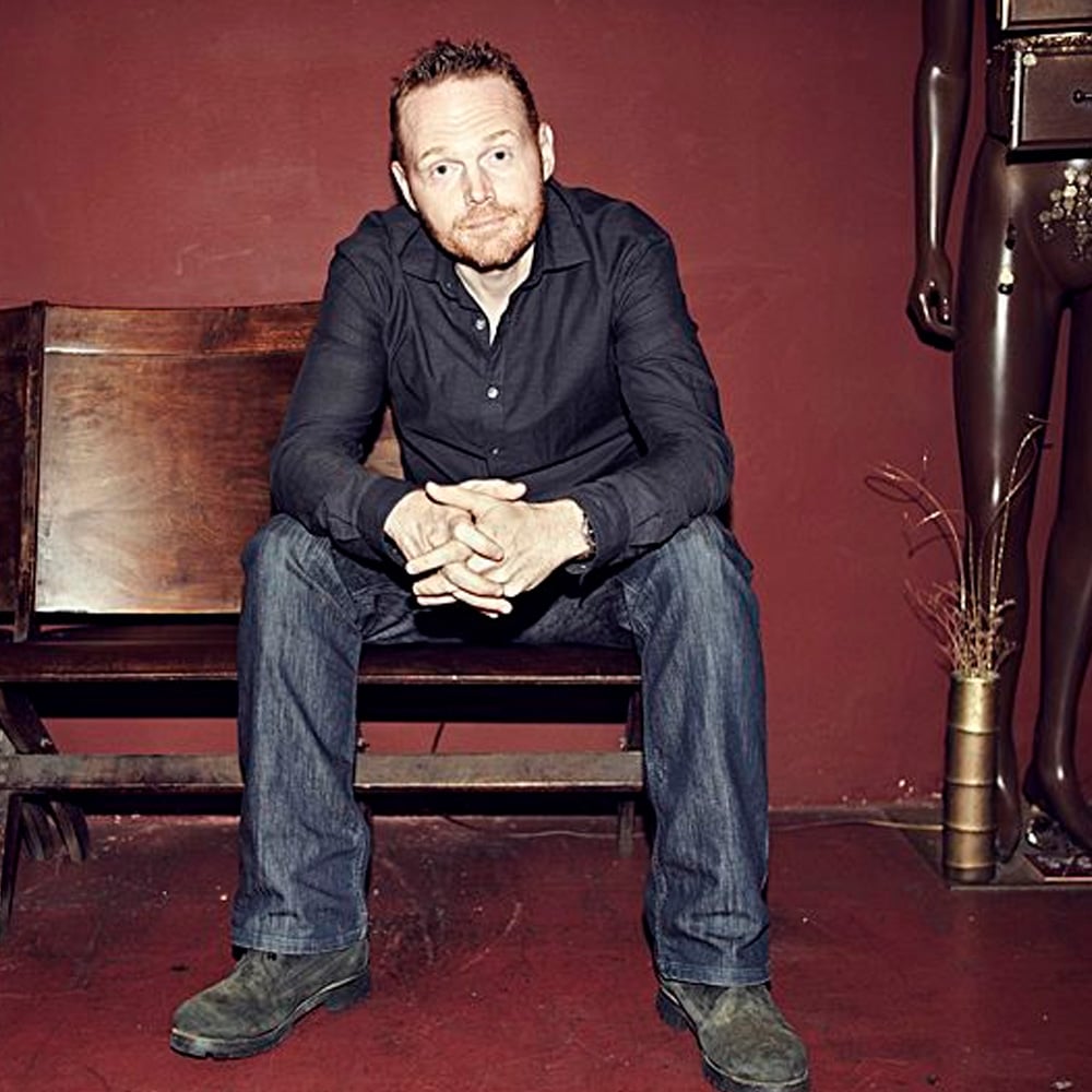 A man with short red hair and a beard sits on a wooden bench against a red wall, wearing a dark shirt and jeans. To the right, part of a mannequin and some decorative branches are visible.