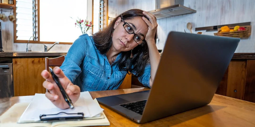 A woman sits at a kitchen table, looking stressed as she holds her head with one hand and a phone with the other, while working on a laptop and taking notes on a clipboard.