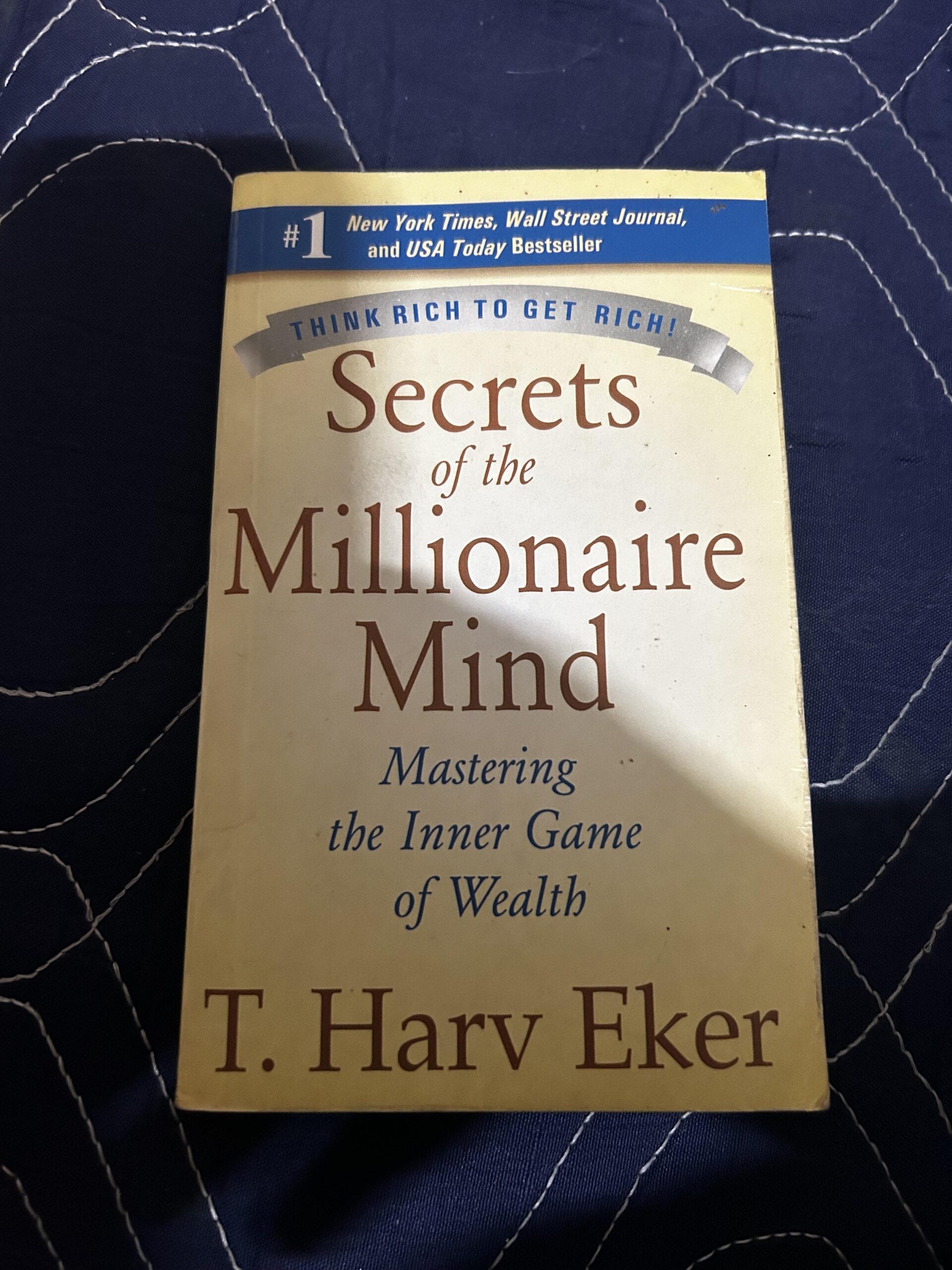 The book "Secrets of the Millionaire Mind" by T. Harv Eker is placed on a dark quilted surface. The cover mentions it's a bestseller and features the subtitle "Mastering the Inner Game of Wealth.