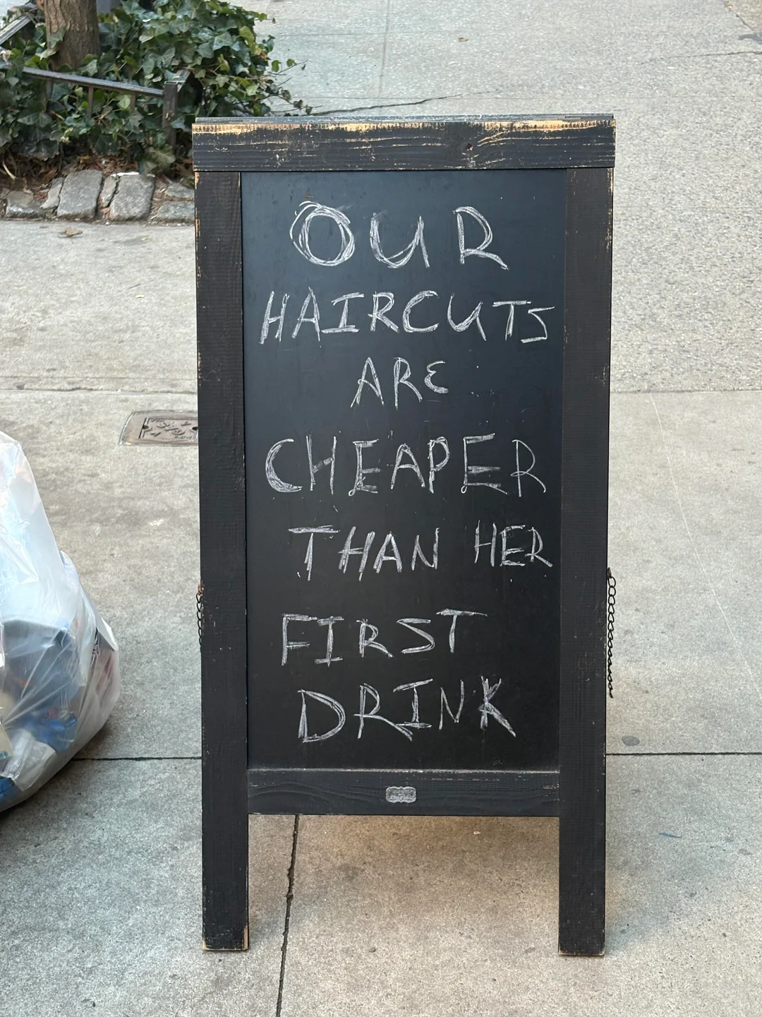A sidewalk chalkboard sign reads: "Our haircuts are cheaper than her first drink." The sign is placed on a concrete sidewalk near a trash bag and some greenery.