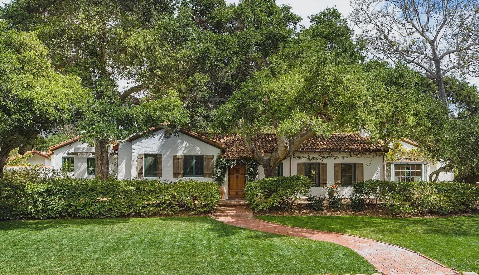 A white stucco house with a red tile roof is surrounded by lush green trees and bushes. A curved brick pathway leads across a well-manicured lawn to the wooden front door.