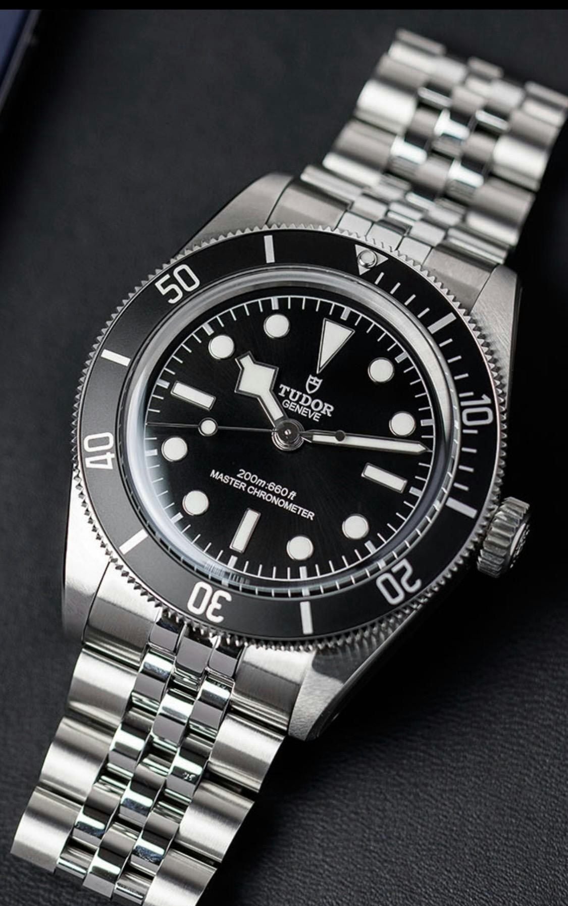 A close-up of a Tudor dive watch with a black dial, silver hour markers, rotating bezel, and stainless steel bracelet, displayed at an angle on a dark surface.