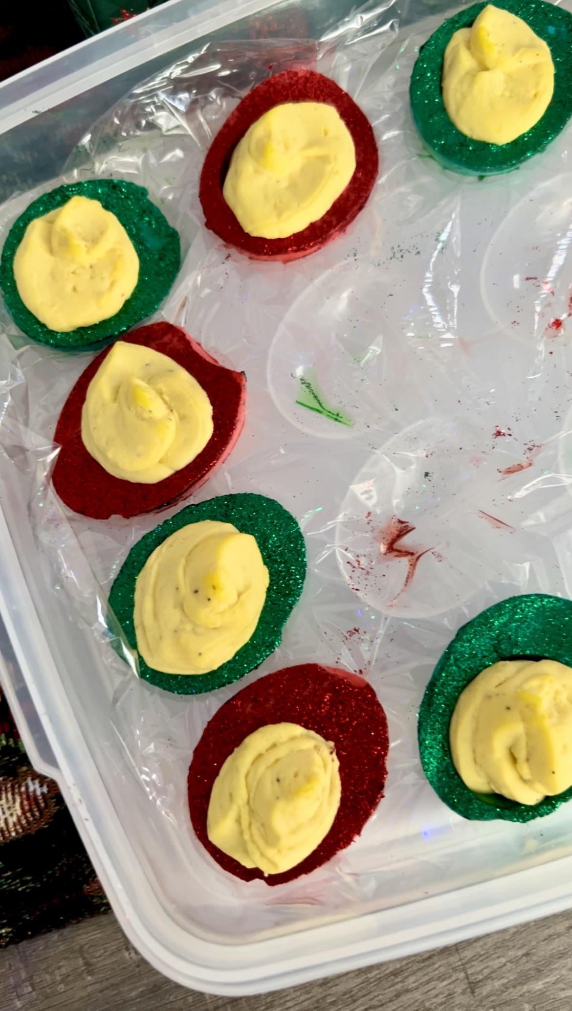 Deviled eggs with yellow filling are placed on red and green glittery bases in a plastic container, giving a festive, holiday-themed appearance.