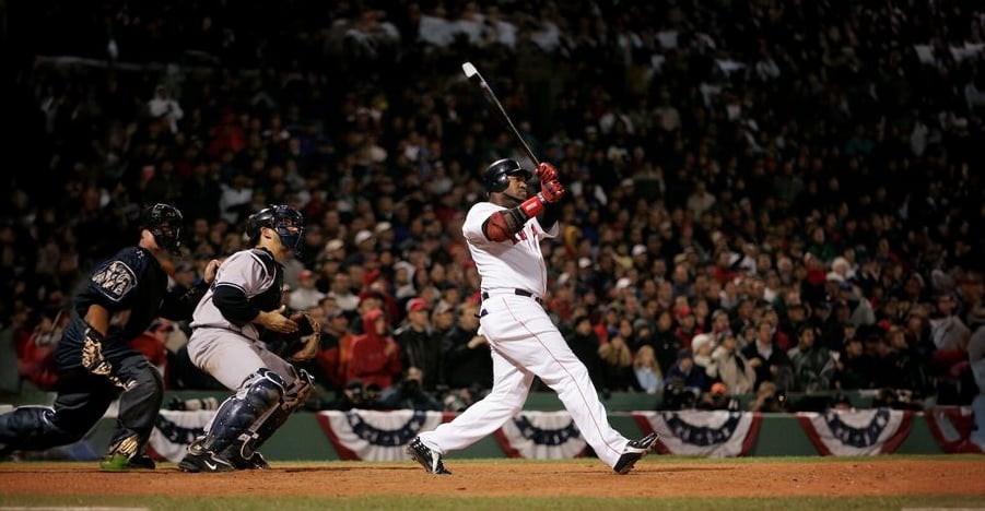 A baseball player in a white uniform swings his bat after hitting the ball, while the catcher and umpire watch. The stadium is filled with spectators and festive banners decorate the stands.
