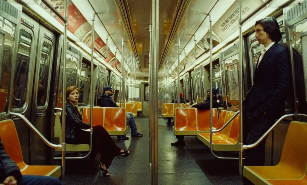 A woman in a black outfit sits on an orange subway seat, looking forward, while a man in a suit stands across from her. The subway car is mostly empty, with a few other passengers seated in the background.