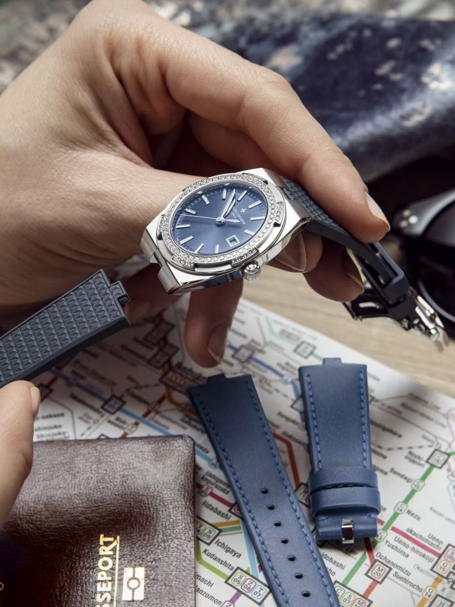 A hand holds a silver and blue wristwatch with interchangeable blue straps above a city map, alongside a brown passport holder and sunglasses.