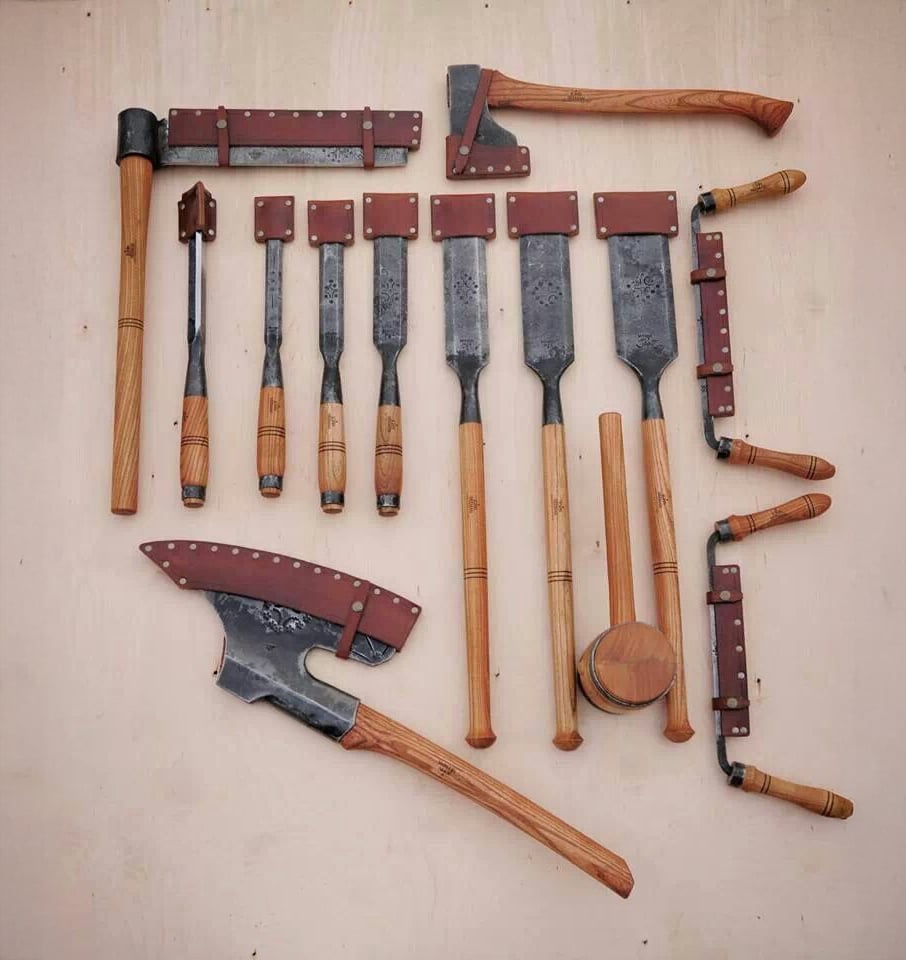 A set of various woodworking tools with wooden handles and metal blades, including saws, axes, chisels, and mallets, neatly arranged on a light-colored surface.
