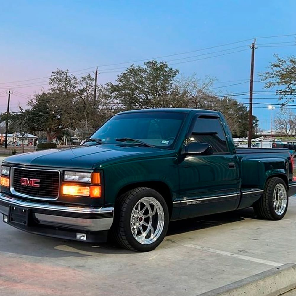 A dark green GMC pickup truck with chrome wheels is parked on a concrete surface. Trees, power lines, and a clear evening sky are visible in the background.