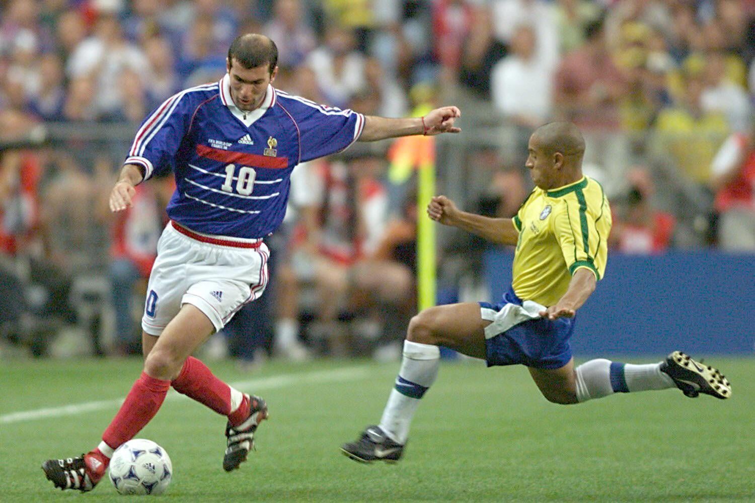 A French soccer player in a blue jersey dribbles the ball while a Brazilian player in a yellow jersey slides in to tackle during a match on a grassy field. Spectators are visible in the blurred background.
