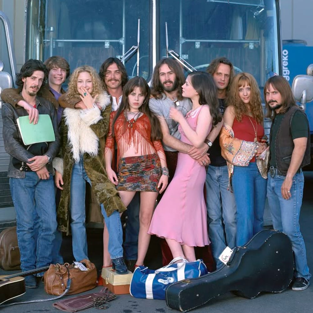 A group of young adults in 1970s-style clothing stand in front of a tour bus with suitcases and guitar cases, smiling and posing together.