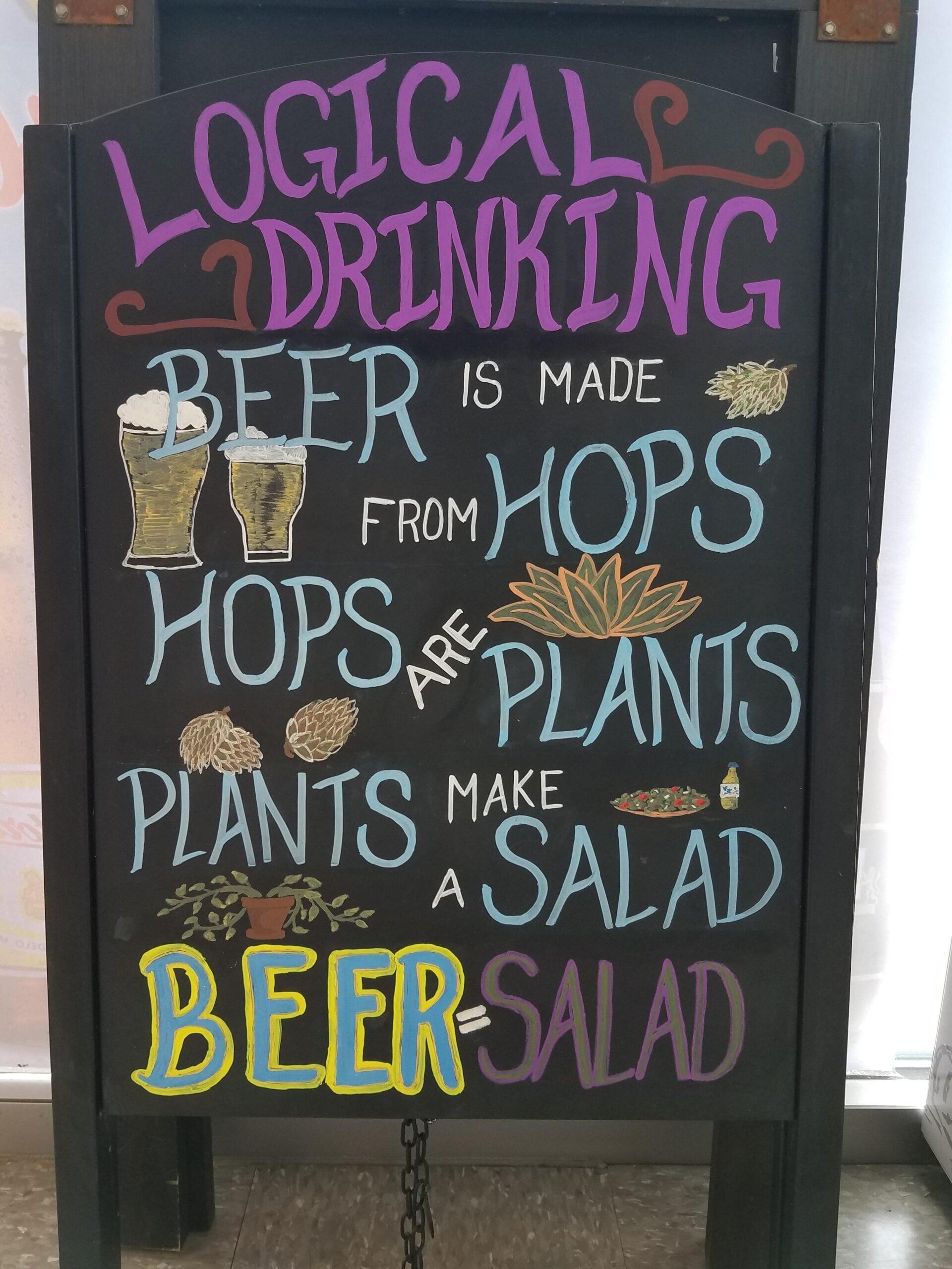 A chalkboard sign reads: "LOGICAL DRINKING. Beer is made from hops. Hops are plants. Plants make a salad. Beer = Salad." The text is decorated with drawings of beer glasses, hops, salad, and plants.