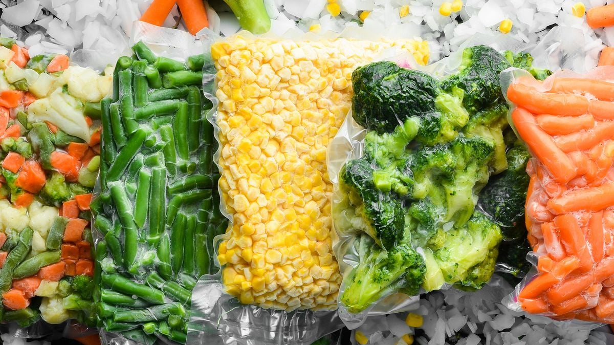Vacuum-sealed bags of frozen vegetables, including mixed vegetables, green beans, corn, broccoli, and baby carrots, are arranged side by side on a bed of ice and scattered vegetables.