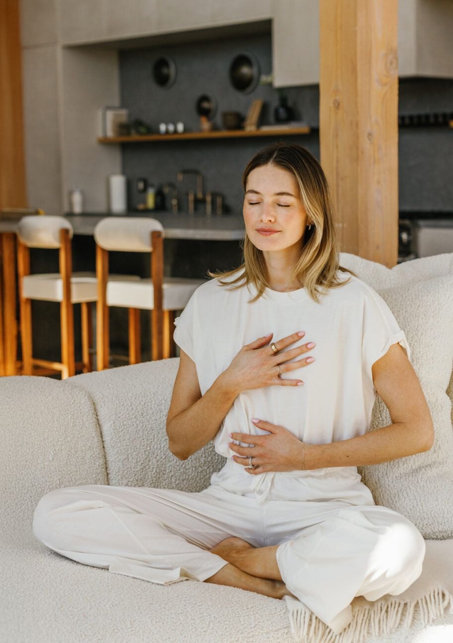 A woman in white clothing sits cross-legged on a couch, eyes closed, with one hand on her chest and the other on her abdomen, appearing calm and peaceful in a modern living room.