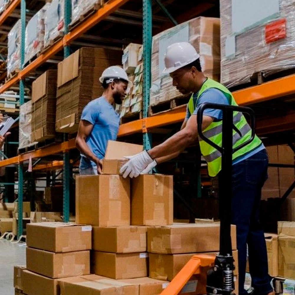 Two workers wearing safety helmets and gloves are stacking cardboard boxes in a warehouse. One wears a safety vest and uses a pallet jack, while shelves filled with boxes are visible in the background.