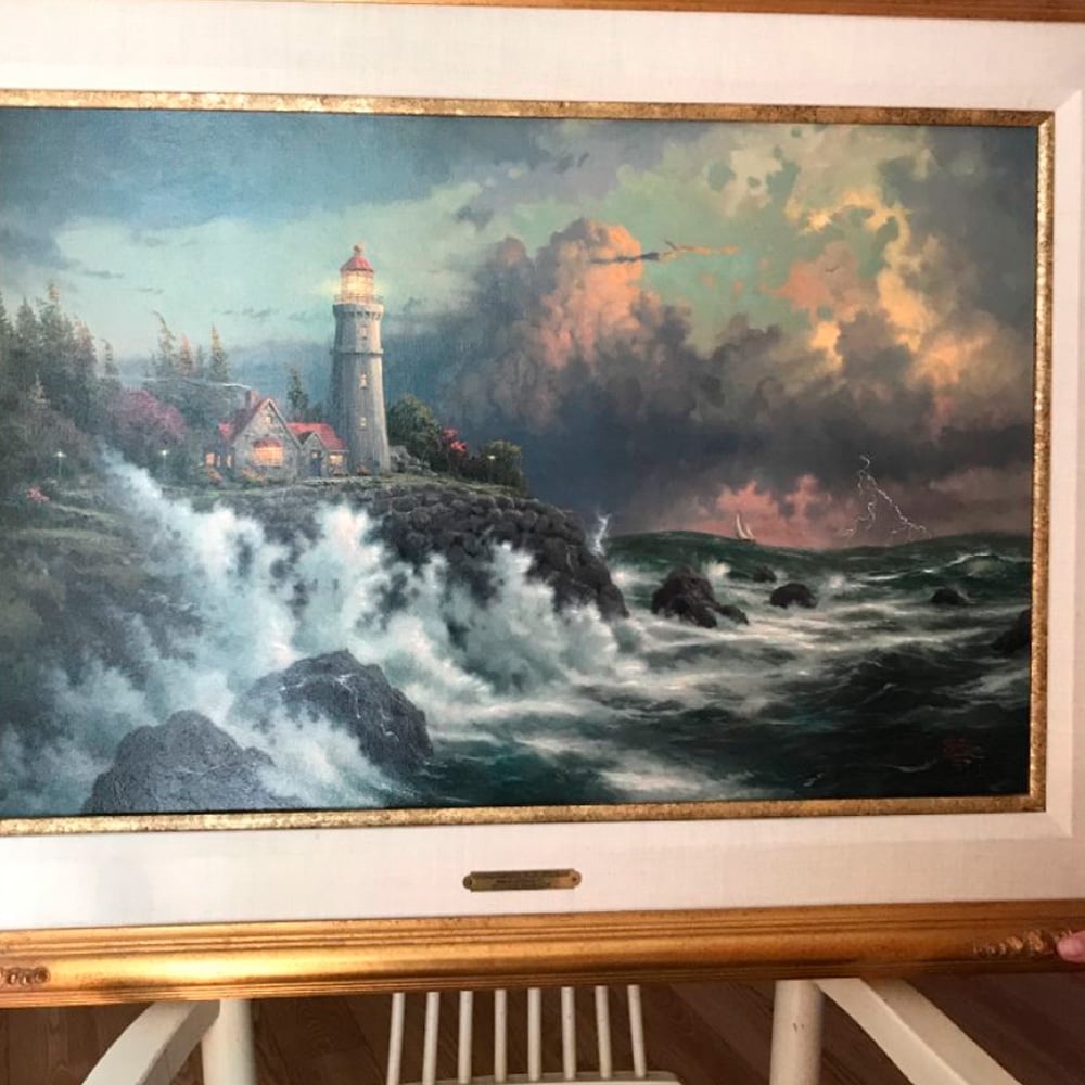 A framed painting depicts crashing waves against rocky cliffs, a lighthouse with a red roof, and a nearby house surrounded by trees. Dramatic clouds and lightning fill the stormy sky above the turbulent sea.