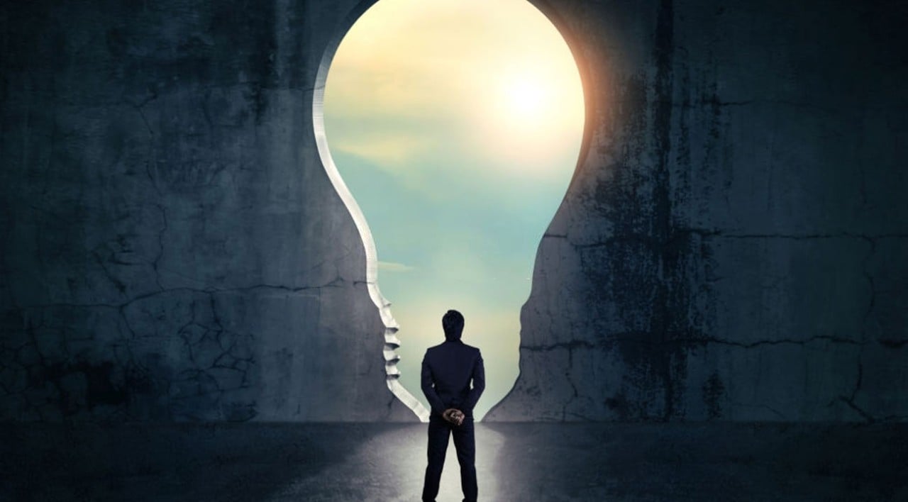 A person in a suit stands in front of a large keyhole-shaped opening in a dark wall, shaped like a light bulb, with bright sunlight and a sky visible through the opening.