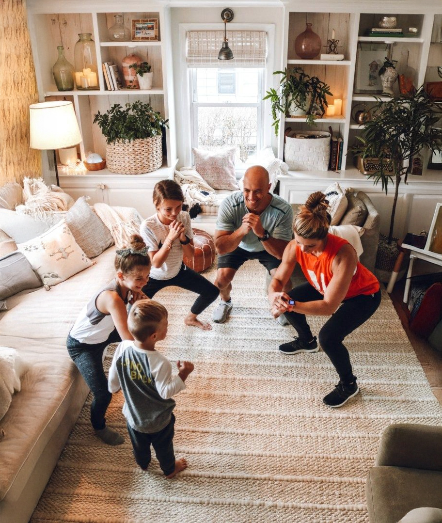 A family of four, two adults and two children, exercise together doing squats in a cozy, well-lit living room with plants, shelves, and soft furnishings. Everyone appears engaged and active.
