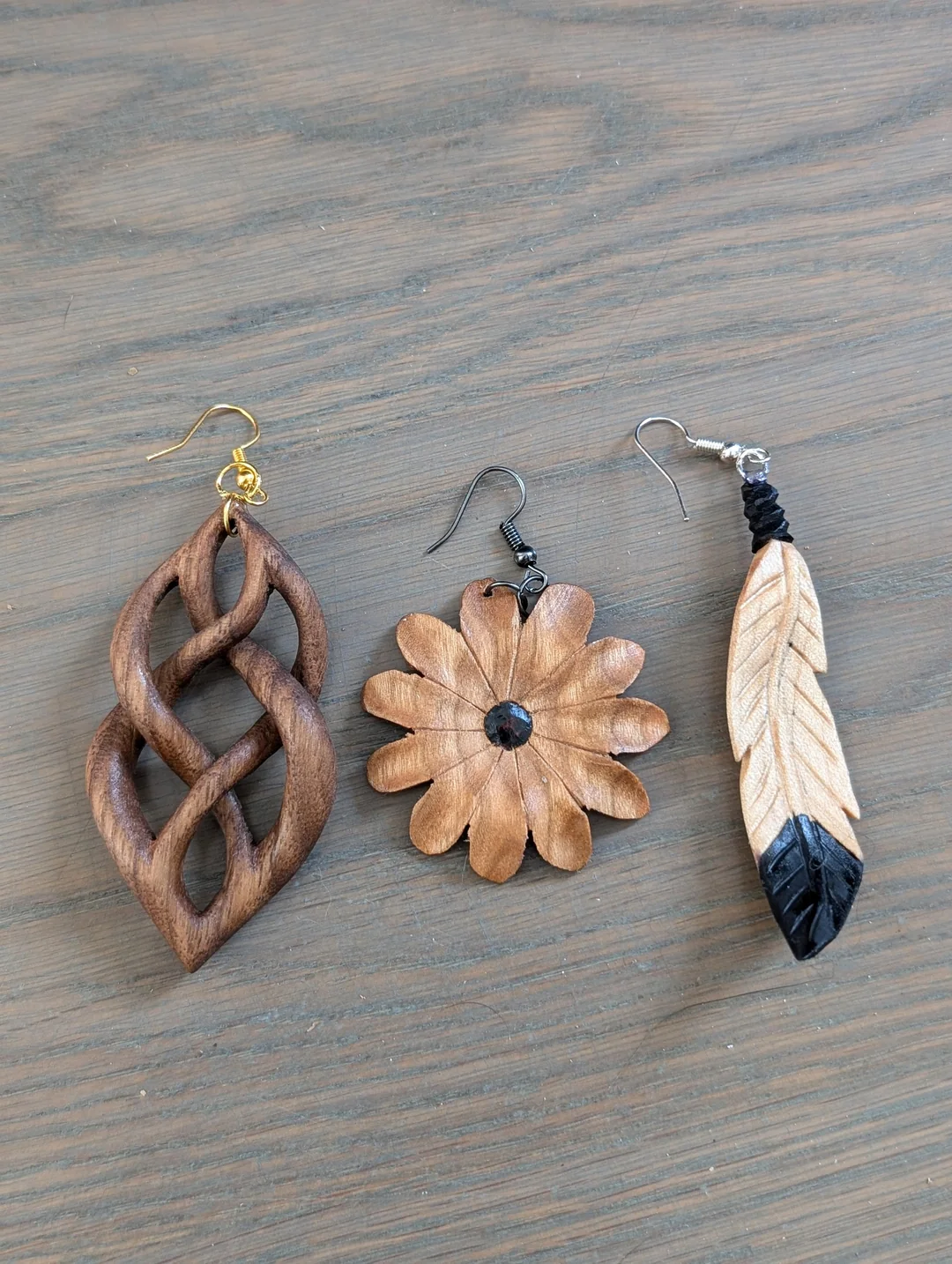 Three wooden earrings on a gray wood surface: a twisted abstract shape, a flower with a black center, and a carved feather painted black at the tip. Each earring has a different color metal hook.