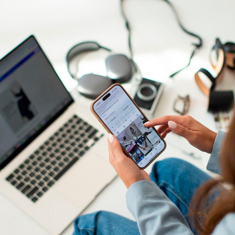 A person uses a smartphone in front of an open laptop, with headphones, a camera, sunglasses, and a watch on a white desk. The phone screen displays a social media app with photos.