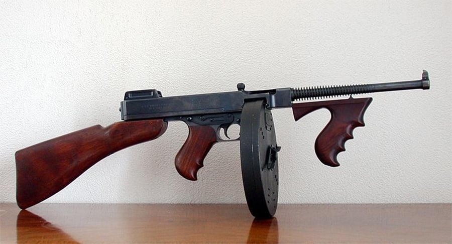 A Thompson submachine gun with a wooden stock, wooden foregrip, and a large drum magazine rests on a wooden surface against a plain white background.