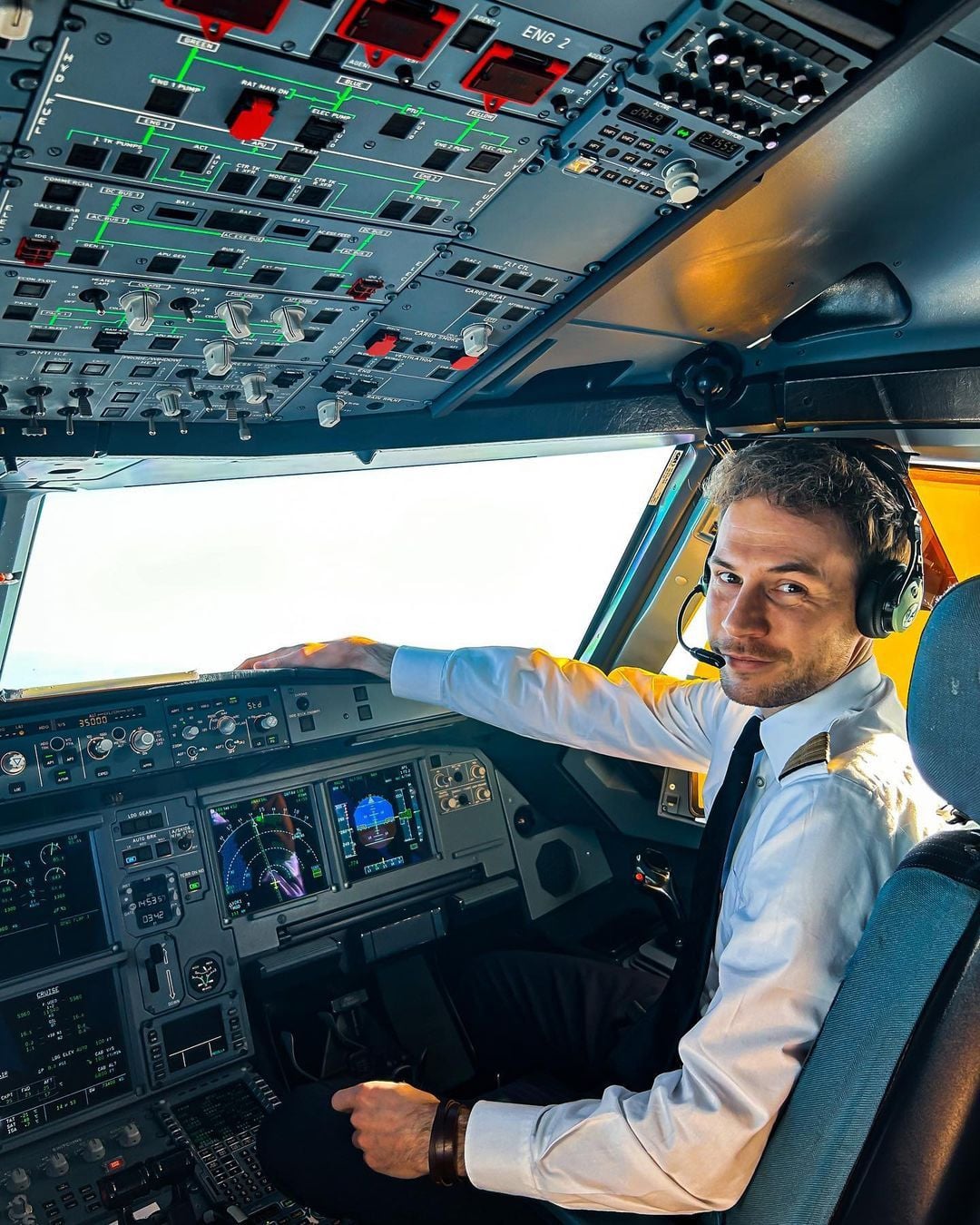 A pilot in uniform sits in an airplane cockpit, one hand on the controls and wearing a headset, looking back and smiling, with various control panels and illuminated buttons visible above and in front of him.