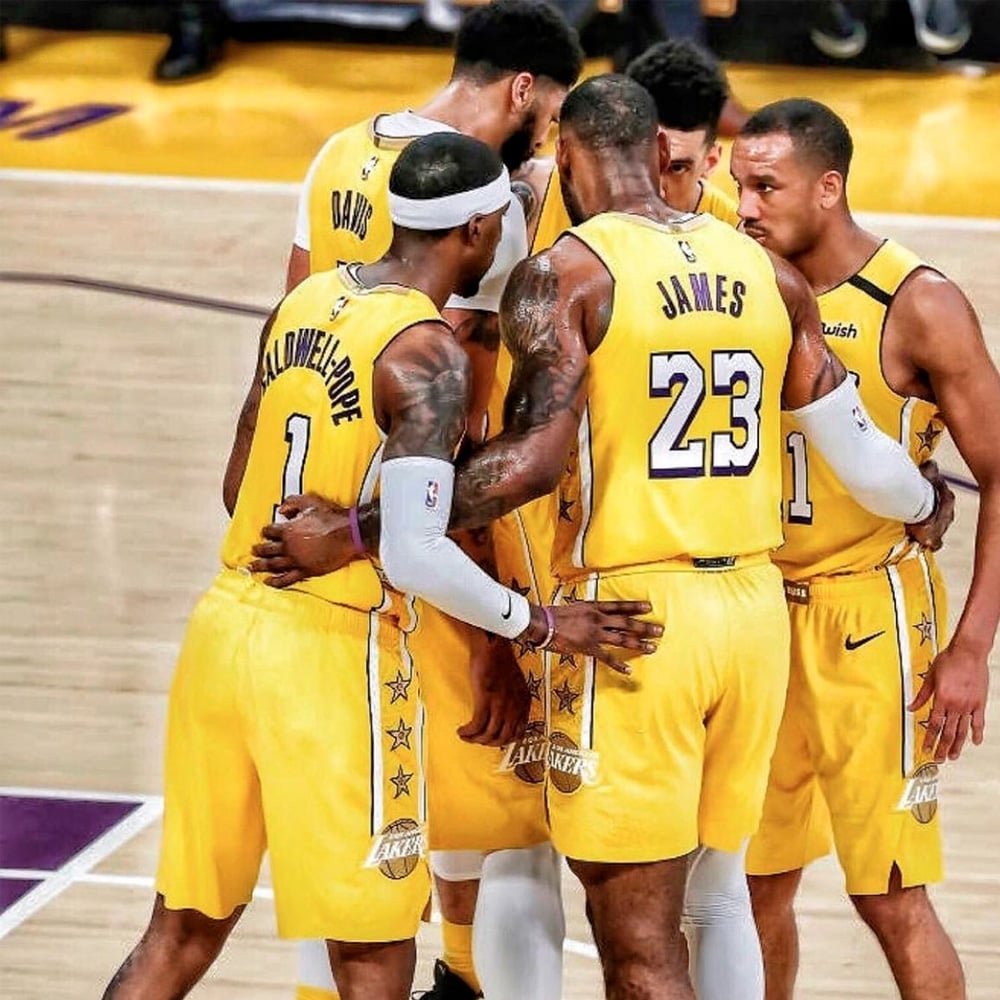 Five basketball players in yellow Los Angeles Lakers uniforms huddle together on the court, displaying teamwork and focus during a game. The players have their arms around each other, preparing for play.