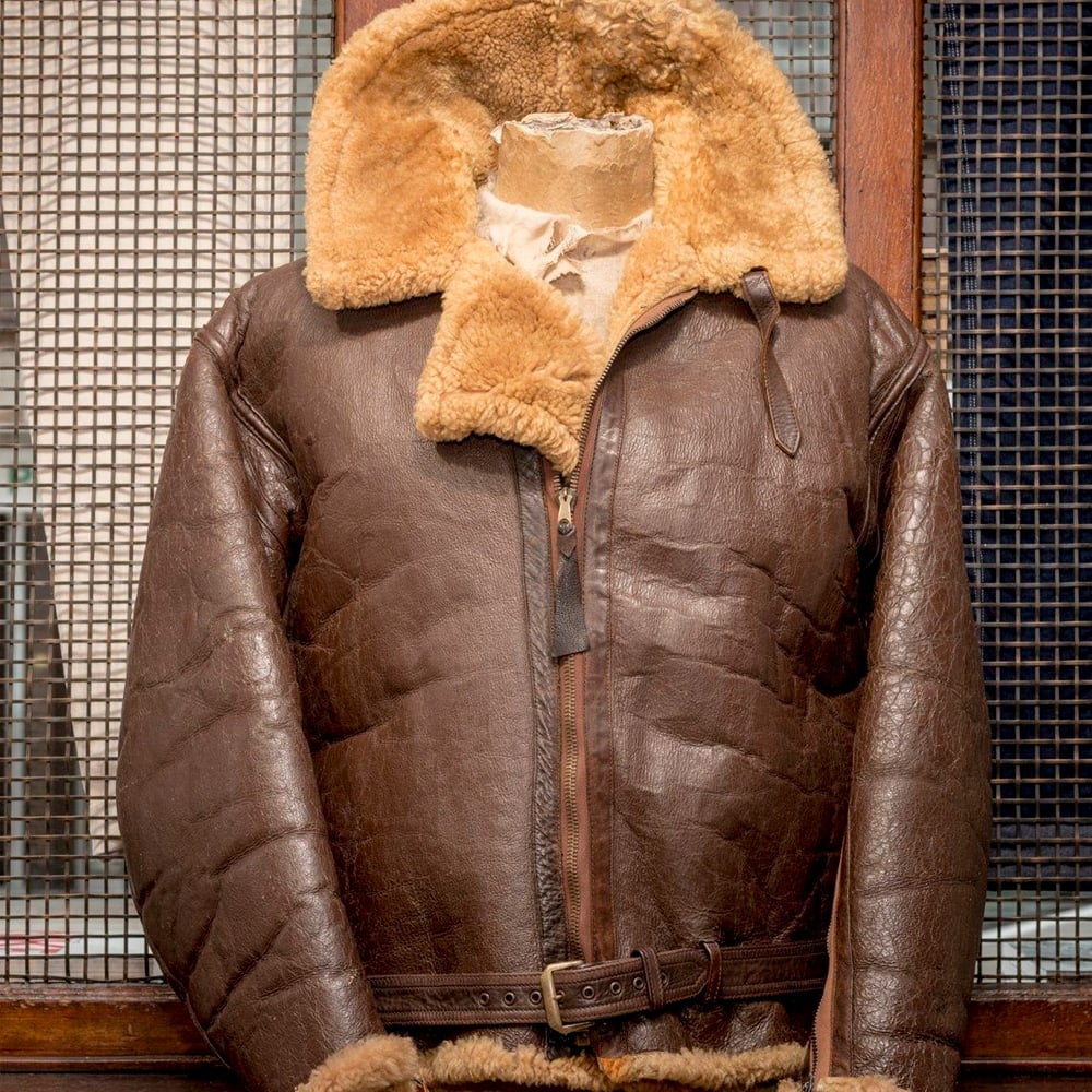 A vintage brown leather bomber jacket with a wide tan shearling collar and cuffs, displayed on a mannequin in front of a metal grid and wooden background.