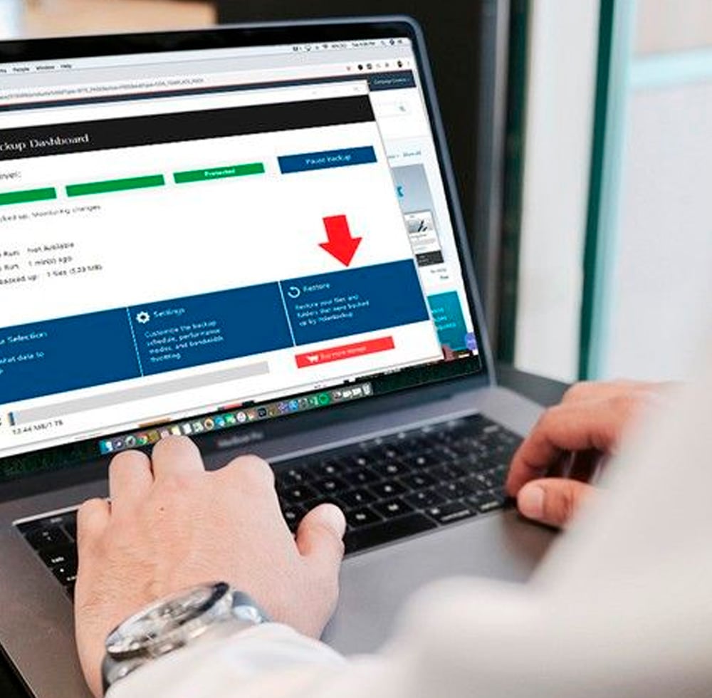 A person uses a laptop displaying a dashboard with tabs and a red arrow pointing to a "Restore" button on the screen. The user's hands are on the keyboard.