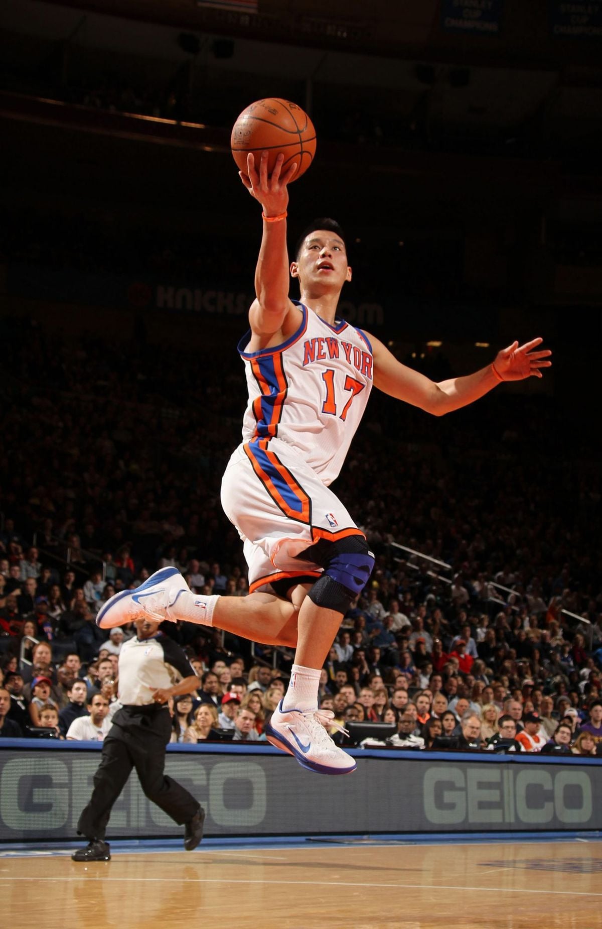 A basketball player wearing a New York Knicks jersey with the number 17 leaps towards the basket for a layup during a game, with fans and a referee visible in the background.