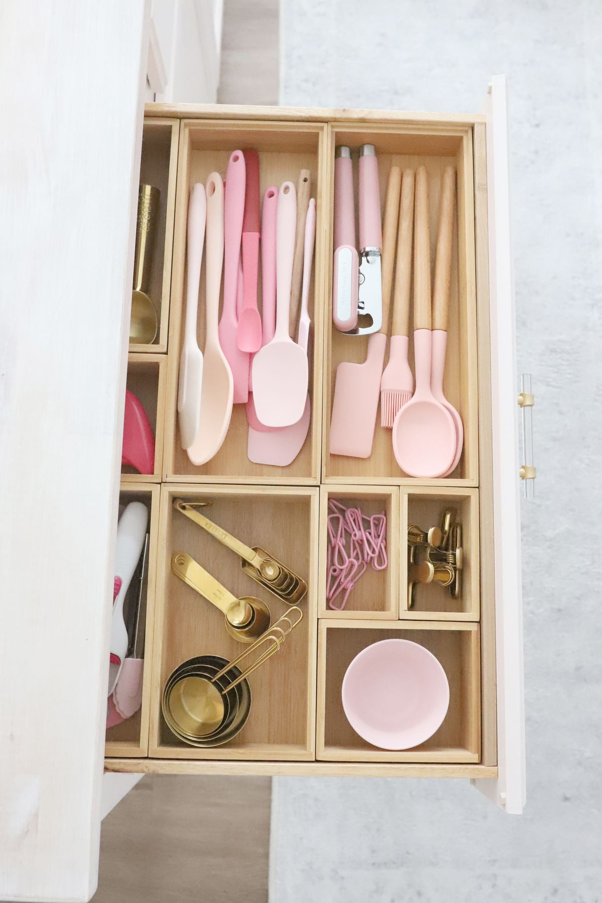 An organized kitchen drawer with pink and wooden utensils, measuring cups and spoons, a can opener, clips, a brush, and a small pink bowl arranged neatly in a wooden divider.