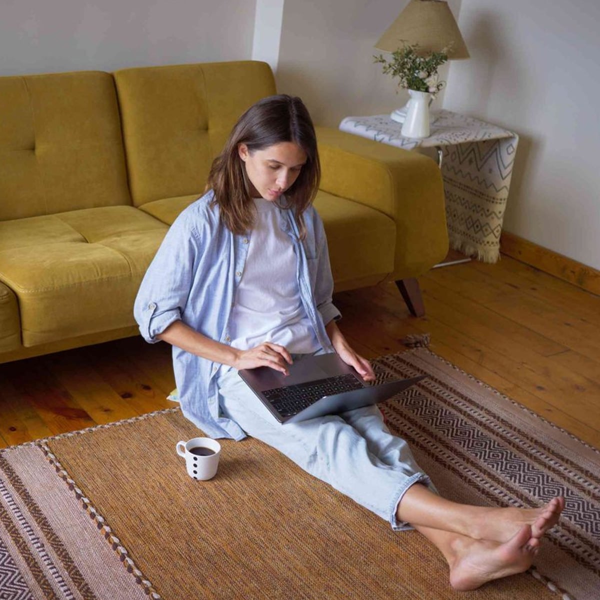 A woman sits on a rug in a living room, using a laptop on her lap. She is barefoot and has a mug beside her. Behind her is a yellow sofa, a side table with a patterned cloth, and a lamp.