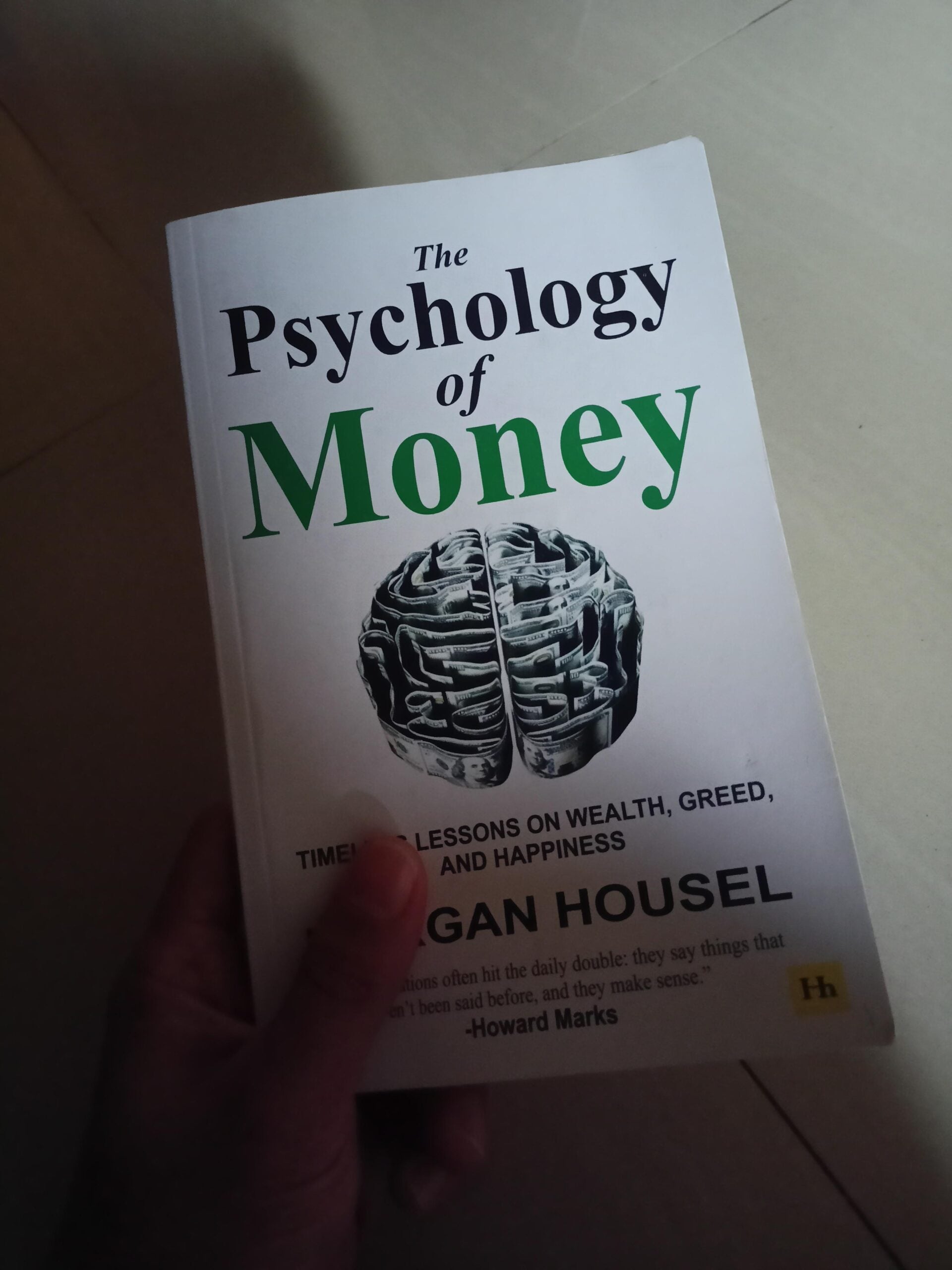 A hand holding the book "The Psychology of Money" by Morgan Housel, featuring a brain made of coins on the cover and the subtitle "Timeless Lessons on Wealth, Greed, and Happiness.