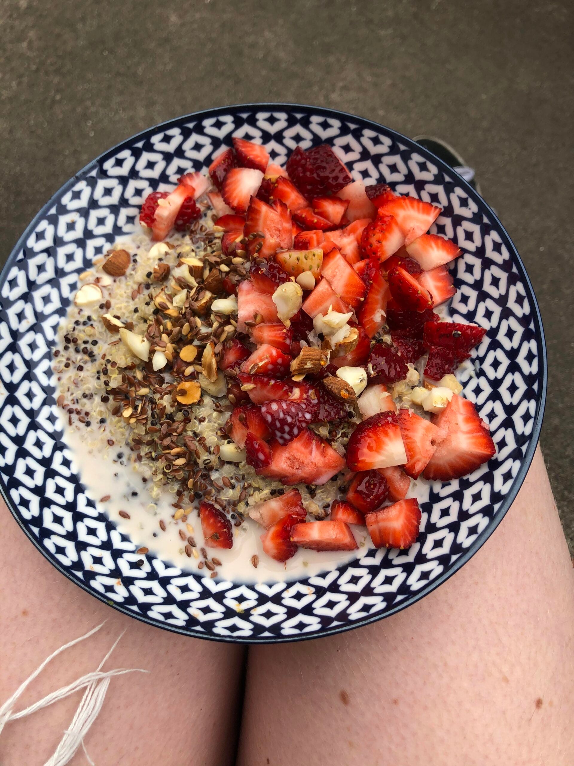 A patterned bowl of oatmeal topped with chopped strawberries, nuts, seeds, and milk, resting on someone's bare legs outdoors.