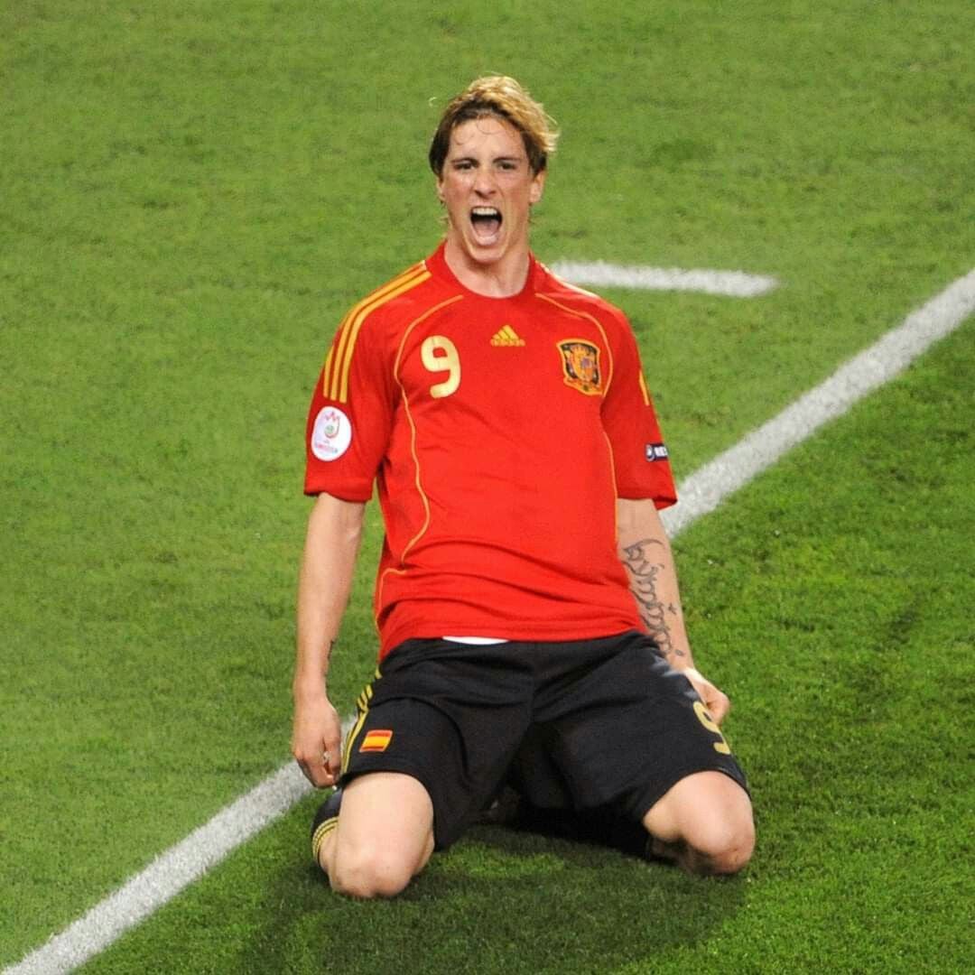 A soccer player in a red Spain jersey with number 9 celebrates on his knees on a grassy field, mouth open in excitement, near a white line on the pitch.