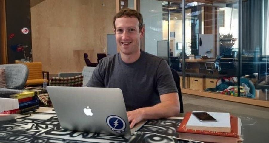 A man with short brown hair, wearing a dark T-shirt, is smiling while sitting at a desk with a laptop in front of him. The desk has books and a phone, and the background shows an office with glass walls.
