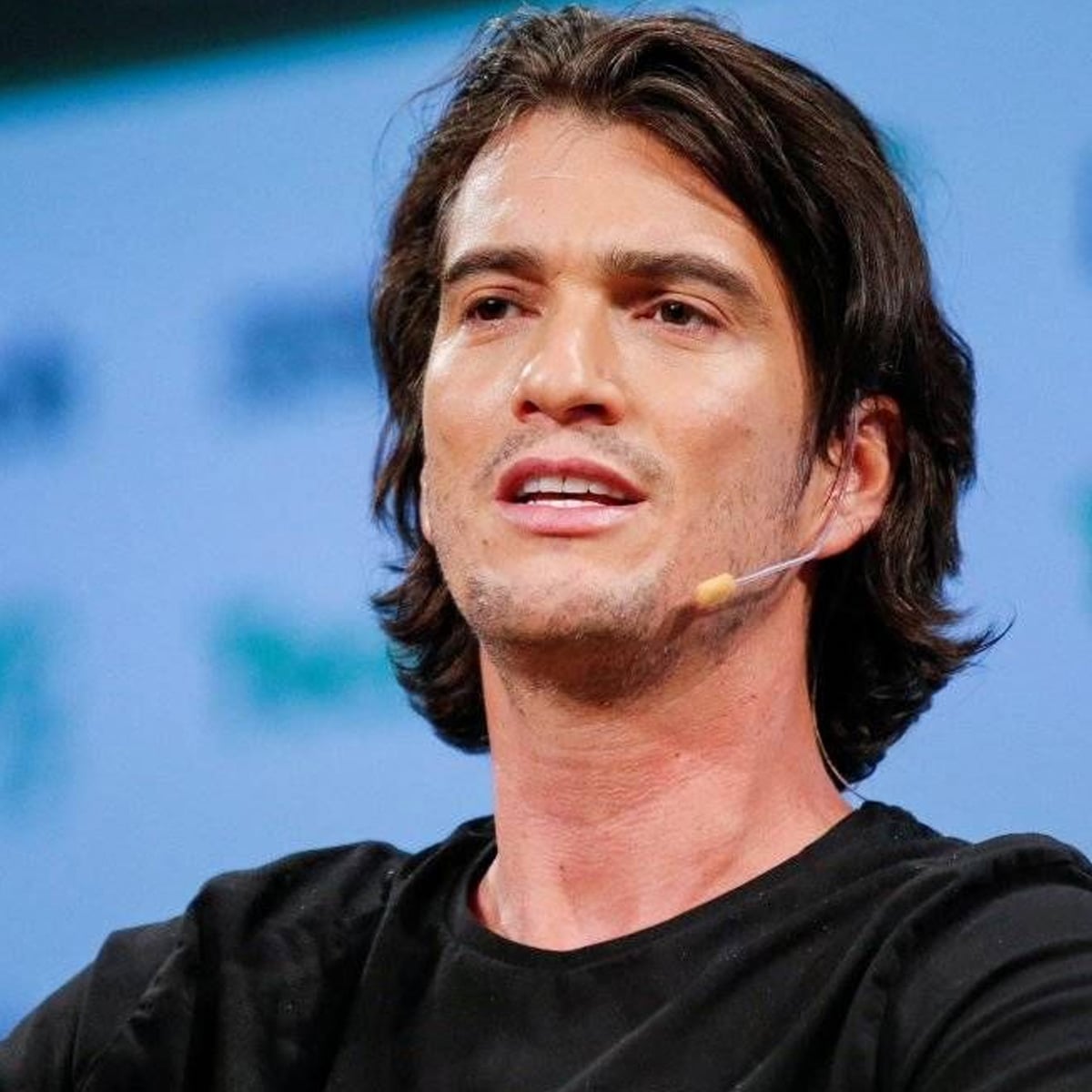 A man with medium-length dark hair wearing a black shirt speaks on stage with a headset microphone, in front of a blurred background with green and blue text.
