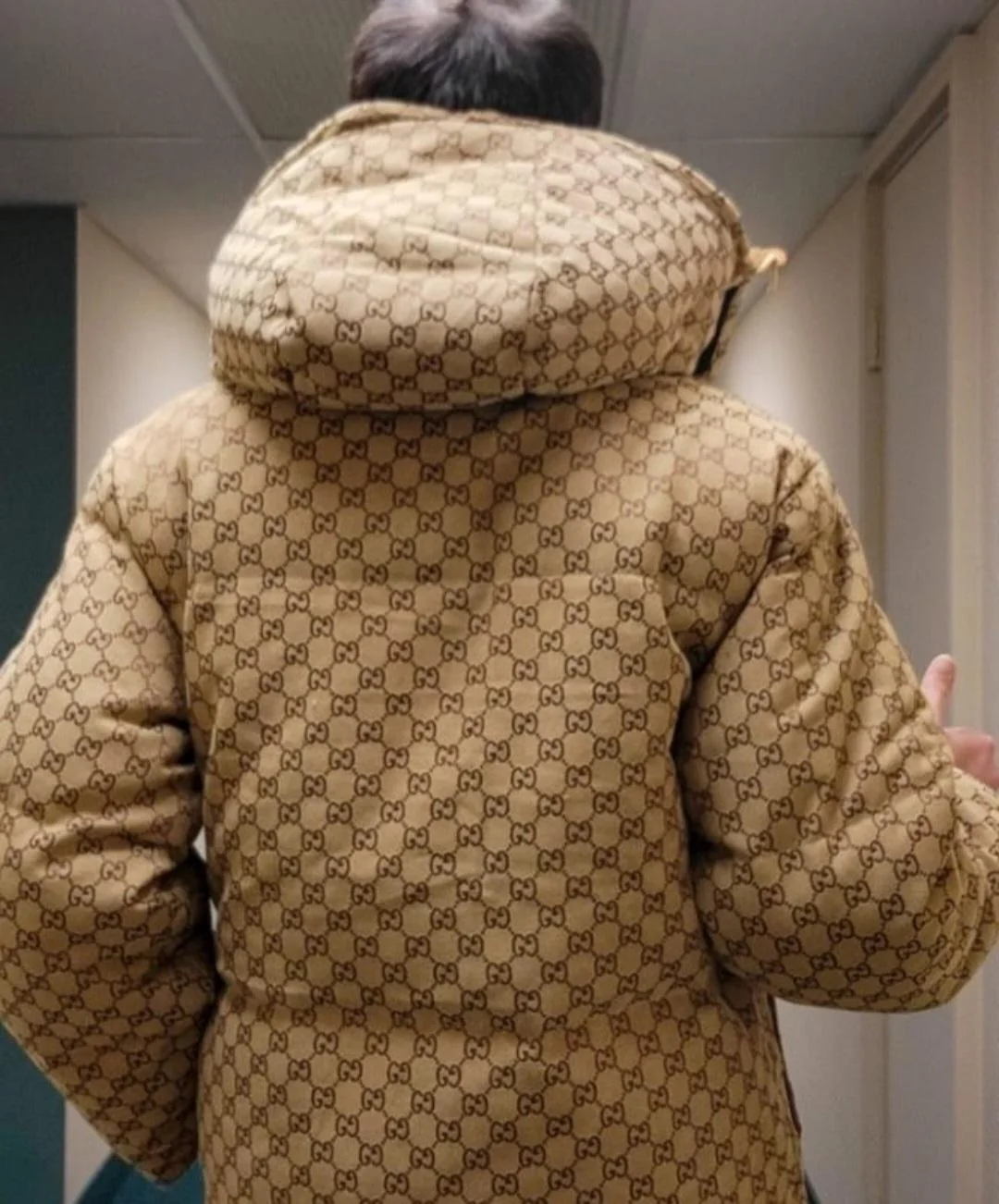 A person is walking down a hallway wearing a tan puffer jacket with a brown Gucci monogram pattern and a hood, seen from behind.