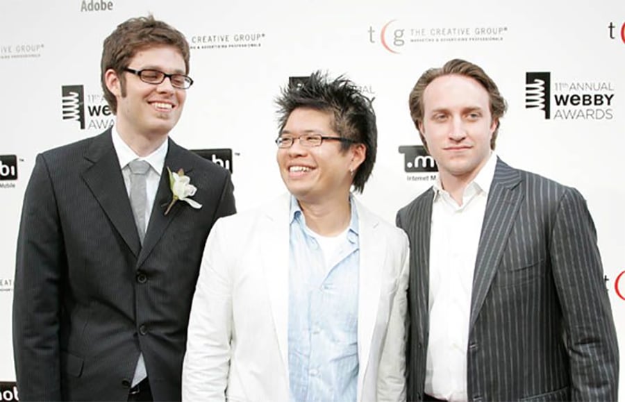 Three men in suits are standing together, smiling, in front of a step-and-repeat banner at the Webby Awards event. Two wear dark suits while the man in the middle wears a white suit and glasses.