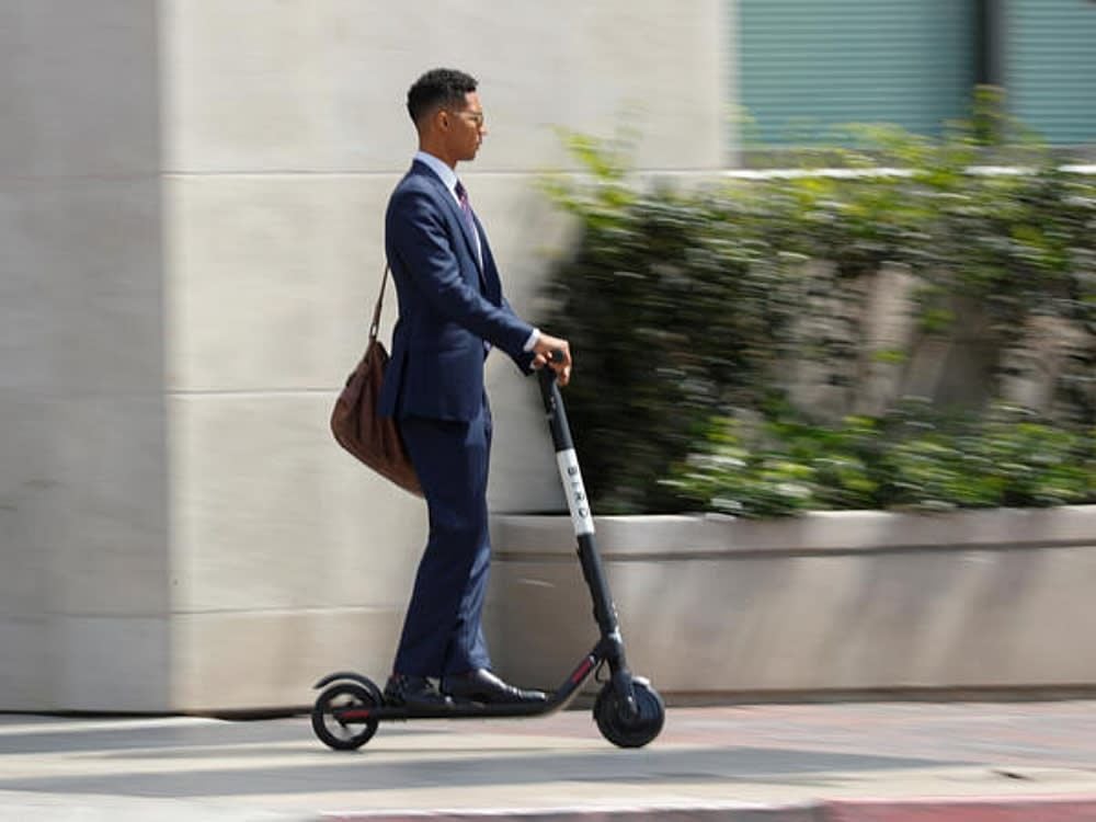 A man in a blue suit and brown shoulder bag rides an electric scooter on a city sidewalk, with blurred greenery and buildings in the background.