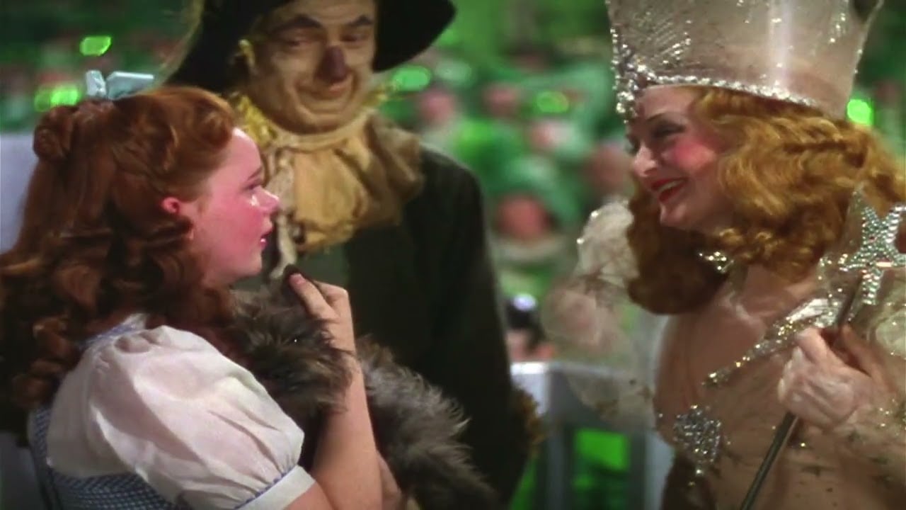 Dorothy, holding Toto, talks to Glinda the Good Witch, who wears a crown and holds a wand; the Scarecrow stands behind them, smiling. The background is green and blurred.