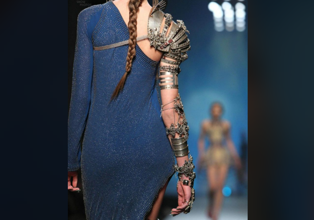 A model walks a runway in a sparkling, sheer blue dress with an asymmetrical back and a single metallic, armored shoulder and arm piece. Another model in a gold outfit is visible blurred in the background.