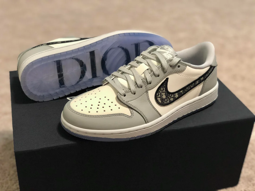 A pair of Nike Air Jordan 1 Low Dior sneakers in white, grey, and black, with the Dior logo on the sole and Dior-branded swoosh, displayed on a black box against a beige background.