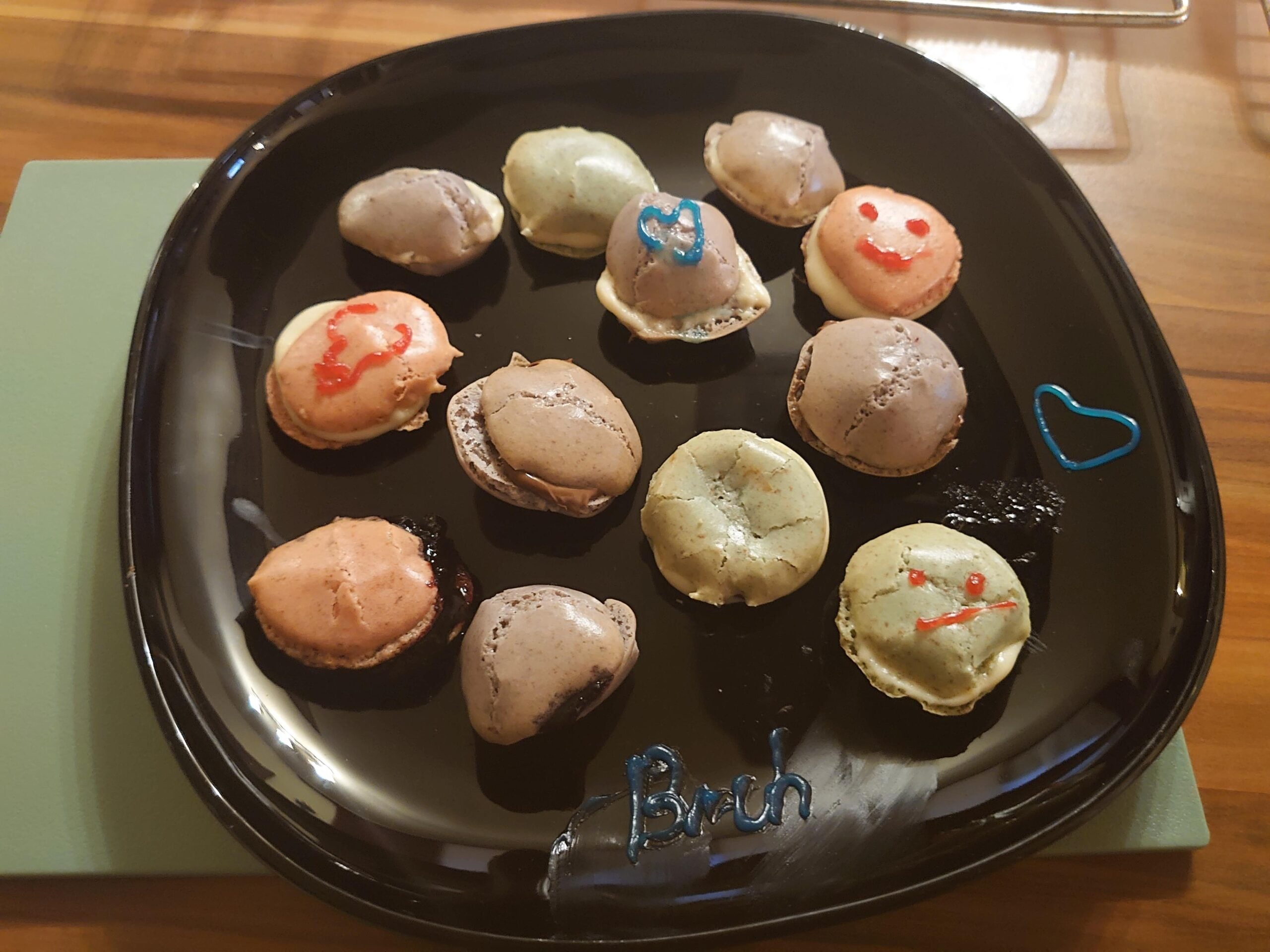 A black plate holds colorful macarons, some decorated with smiley faces and hearts in icing. The word "bruh" is also written in blue icing on the plate. The plate sits on a wooden surface.