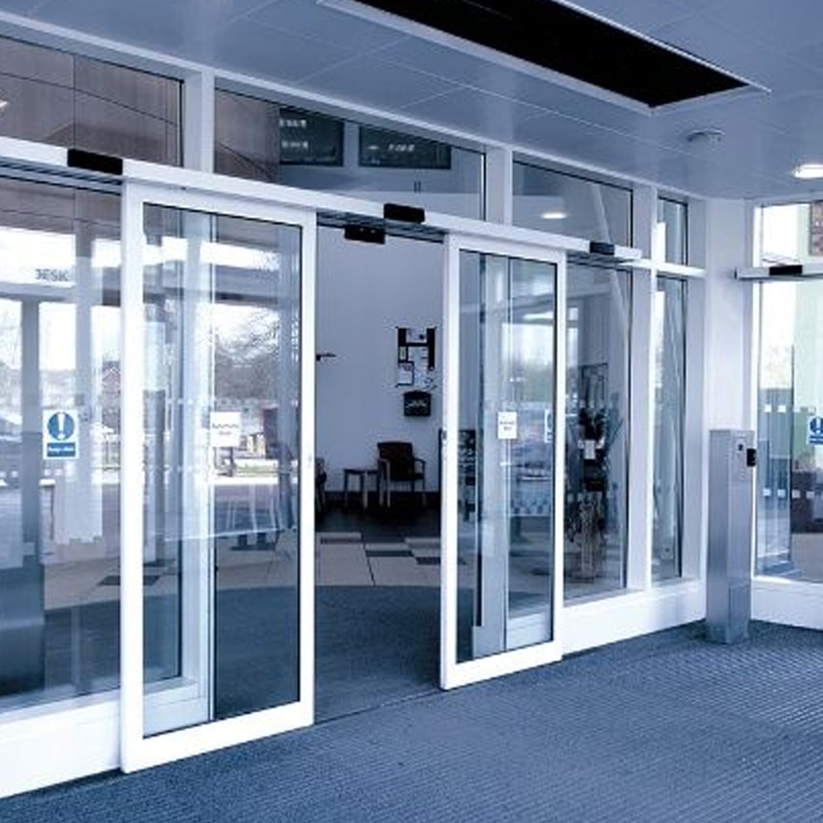 Automatic glass sliding doors open to a modern building entrance, revealing a lobby area with chairs and large windows. The space is well-lit with overhead lights and natural light from outside.